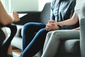 The image focuses on the hands of a couple. They are sat on a sofa holding hands. We cannot see their heads
