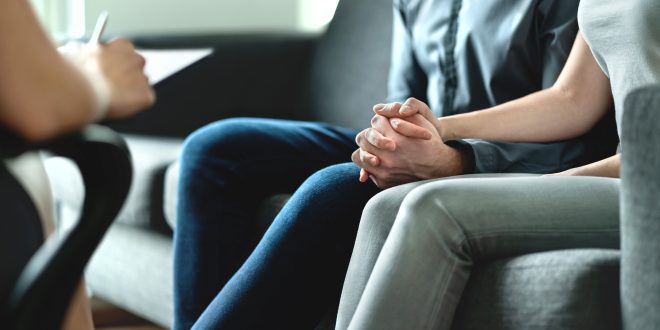 The image focuses on the hands of a couple. They are sat on a sofa holding hands. We cannot see their heads