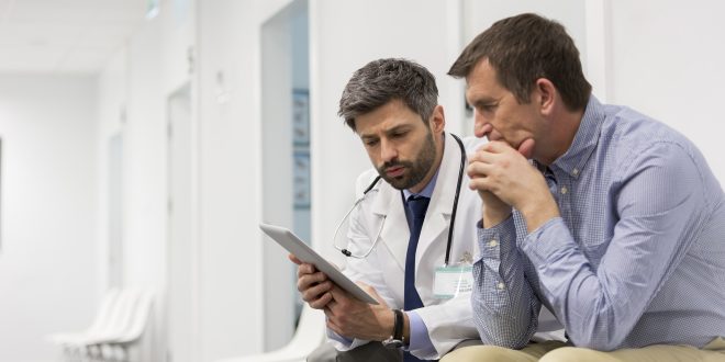 A doctor holding a digital tablet is talking with a middle aged man. They are sitting in a hospital corridor.
