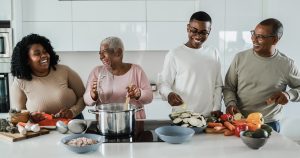 A family group are are cooking together in a modern looking kitchen. From left to right they are a young black woman, she is smiling at an older black woman who is stirring a large cooking pot. A young black man wearing glasses and an older black man wearing glasses are also smiling and helping to prepare colourful vegetables.