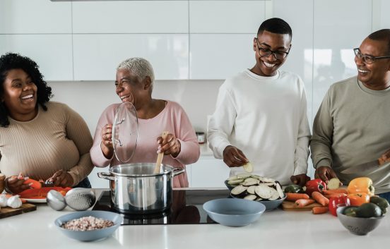 A family group are are cooking together in a modern looking kitchen. From left to right they are a young black woman, she is smiling at an older black woman who is stirring a large cooking pot. A young black man wearing glasses and an older black man wearing glasses are also smiling and helping to prepare colourful vegetables.