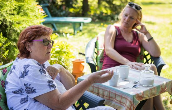 Two middle aged women are having a serious conversation. They are in a garden on a sunny day. There is a small table between them with mugs and teaspoons on it.