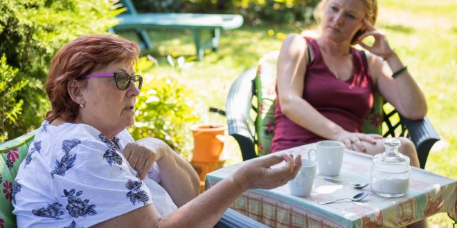 Two middle aged women are having a serious conversation. They are in a garden on a sunny day. There is a small table between them with mugs and teaspoons on it.