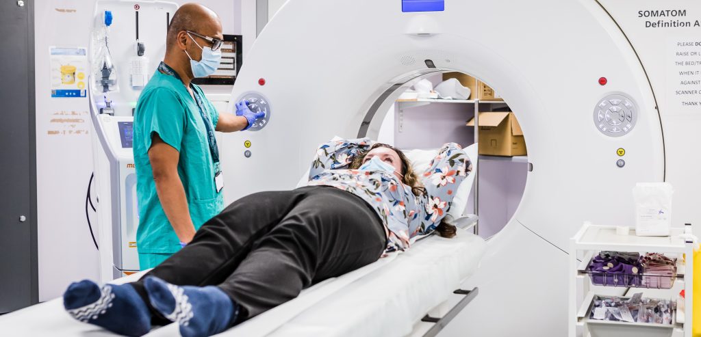 A woman in trousers and a floral shirt is lying on a bed with her arms and head resting on a pillow. The bed is in front of the hole in the middle of a CT scanner. The scanner is a large white ring with several buttons on it. A man in surgical scrubs and a medical face mask is standing next to the woman and pressing some of the buttons on the scanner.