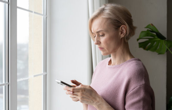 A middle aged woman with a concerned expression is standing beside a window, intently looking at her smartphone. The diffused natural light suggests an overcast day, and she appears to be concerned and is reading information on her phone.