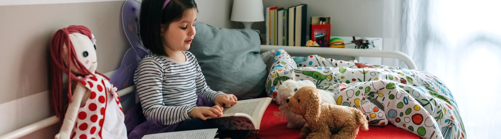 Little girl disguised as a butterfly sitting on the bed reading a book to her toys