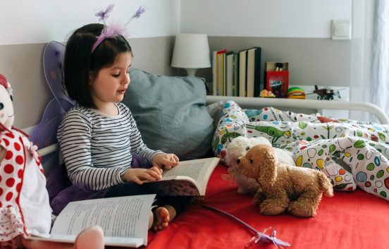 Little girl disguised as a butterfly sitting on the bed reading a book to her toys