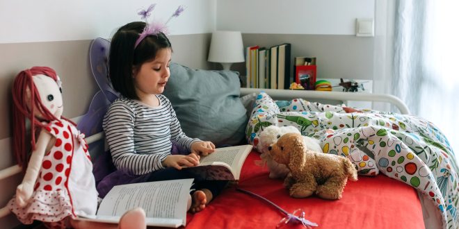 Little girl disguised as a butterfly sitting on the bed reading a book to her toys