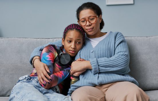 A mum hugs her teenage daughter who looks worried. The are sitting on a sofa.