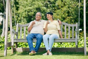 A middle aged man and woman sit on a swing seat outdoors. They both have mugs. The man has his arm around the woman's shoulder. They seem happy and relaxed together.
