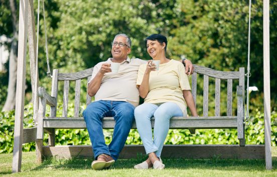 A middle aged man and woman sit on a swing seat outdoors. They both have mugs. The man has his arm around the woman's shoulder. They seem happy and relaxed together.