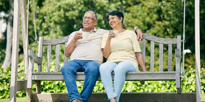 A middle aged man and woman sit on a swing seat outdoors. They both have mugs. The man has his arm around the woman's shoulder. They seem happy and relaxed together.