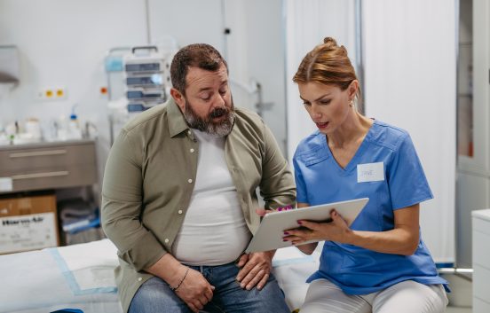 A man sits on a hospital bed. He is talking to a female healthcare professional. She is showing him something on a digital tablet.