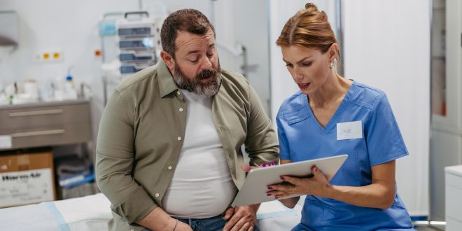 A man sits on a hospital bed. He is talking to a female healthcare professional. She is showing him something on a digital tablet.