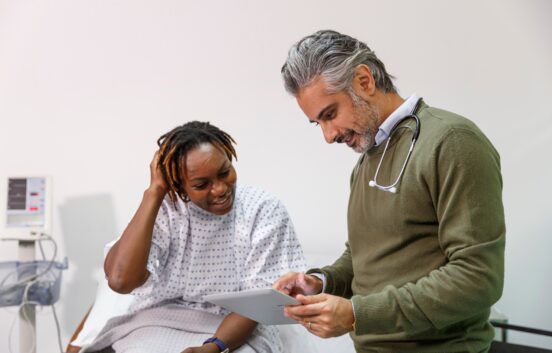 The image shows a middle aged black women wearing a hospital gown. She is sat with a sheet over her legs. A male doctor with greying hair, stethoscope and a khaki jumper is stood next to her. They are both looking at a piece of paper the doctor is holding. They look relaxed and friendly.
