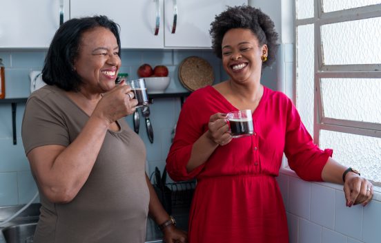 Two black women are drinking coffee from glass mugs. They are smiling and standing in a kitchen next to a window. From their ages they could be mother and daughter.