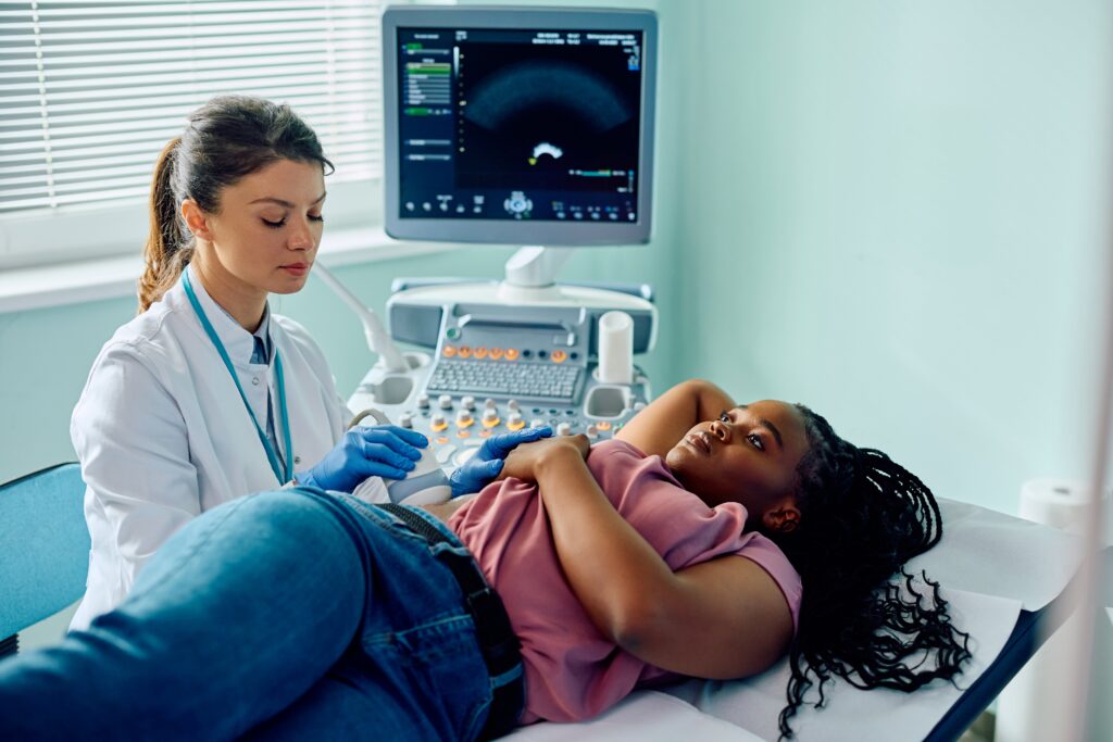 A young woman is lying on a medical bed on her left side. She has slightly lifter her pink T-shirt to expose a small amount of skin just below her ribs on her right side. There is an ultrasound machine in the background. This is being operated by a female medical professional in a white coat. She has placed the probe of the ultrasound on the skin of the woman in the bed, in the space provided by lifting up the T-shirt.