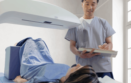 A woman wearing a surgical gown lies on her back, her legs are propped up with a block under her knees. There is a large white scanning arm over her hips. A male healthcare professional wearing scrubs stands next to her he looks friendly and is taking notes.