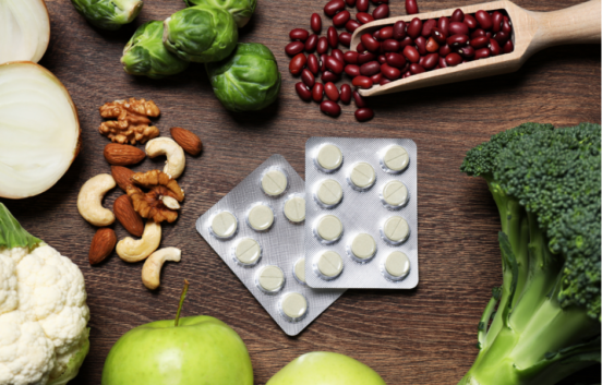 a table with two packets of round white pills in the middle, around them are a variety of healthy foods including fruit, vegetables, nuts and beans.