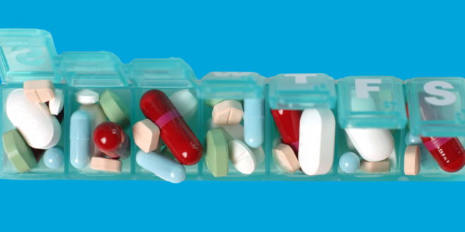 A blue green plastic pill holder with seven compartments. The lids are partly open to show a number of different sized, coloured and shaped pills and capsules in each compartment. The top of the last two lids is visible The first says F the second says S