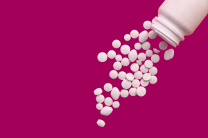 A plain white plastic pill bottle with about 40 different shaped and sized white pills spilling out of it.